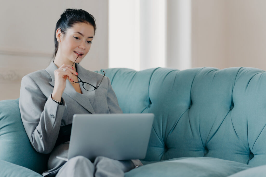 Serious woman entrepreneur takes off glasses, focused in laptop computer screen, analyzes statistics data, looks through financial information, works online, sits on sofa in coworking space.